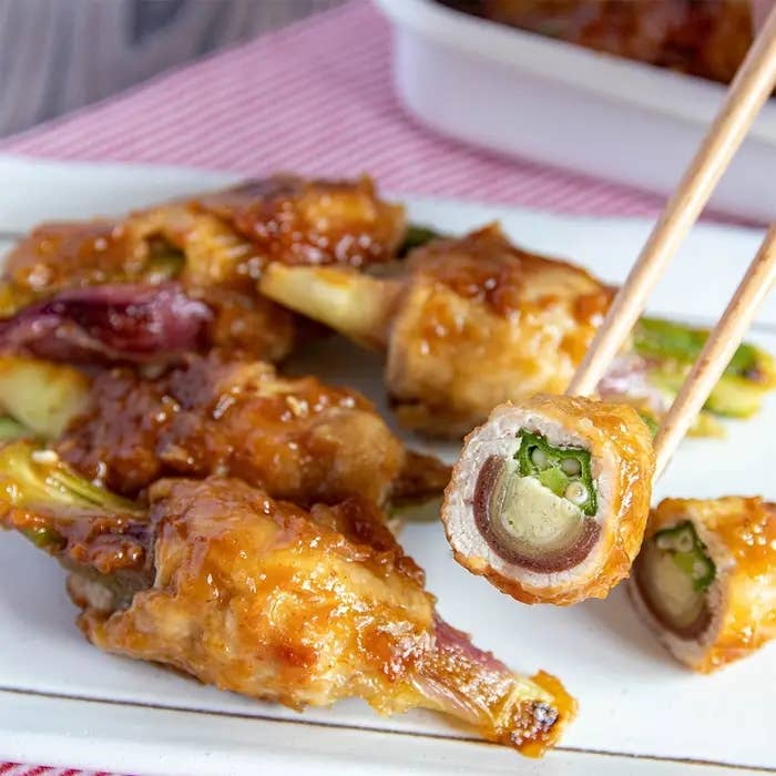 オクラを肉で巻いて焼いた料理が皿に盛り付けられ、箸で1つ持ち上げられている。