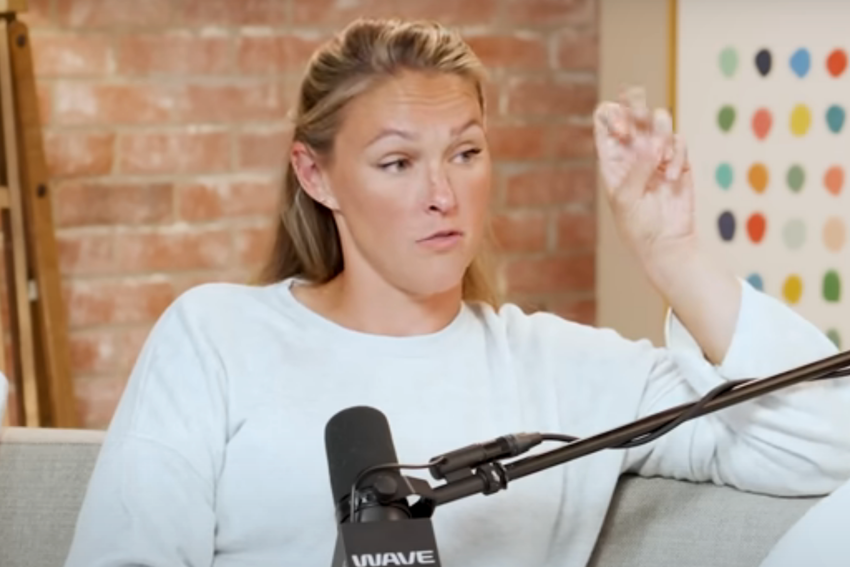 Person speaking into a microphone on a podcast, seated against a brick wall, wearing a casual outfit