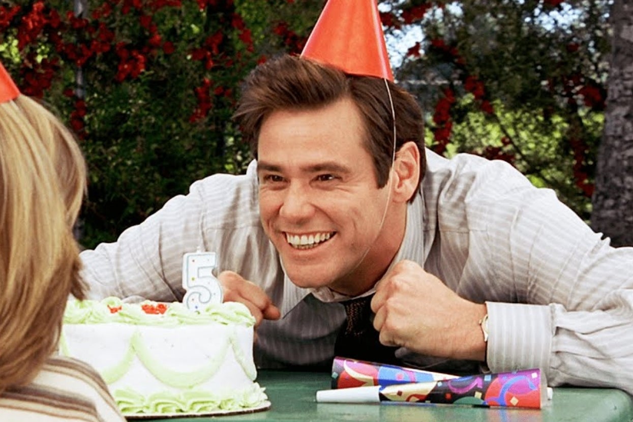 Man in a party hat smiling widely near a cake with a number 5 candle. Festive scene suggests celebration