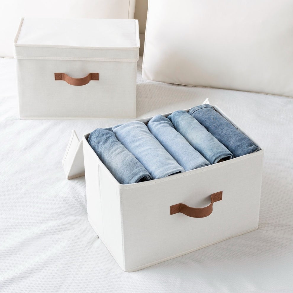 Storage boxes on a bed, one open showing neatly rolled clothes inside. Boxes have leather handles. Featured in a shopping article