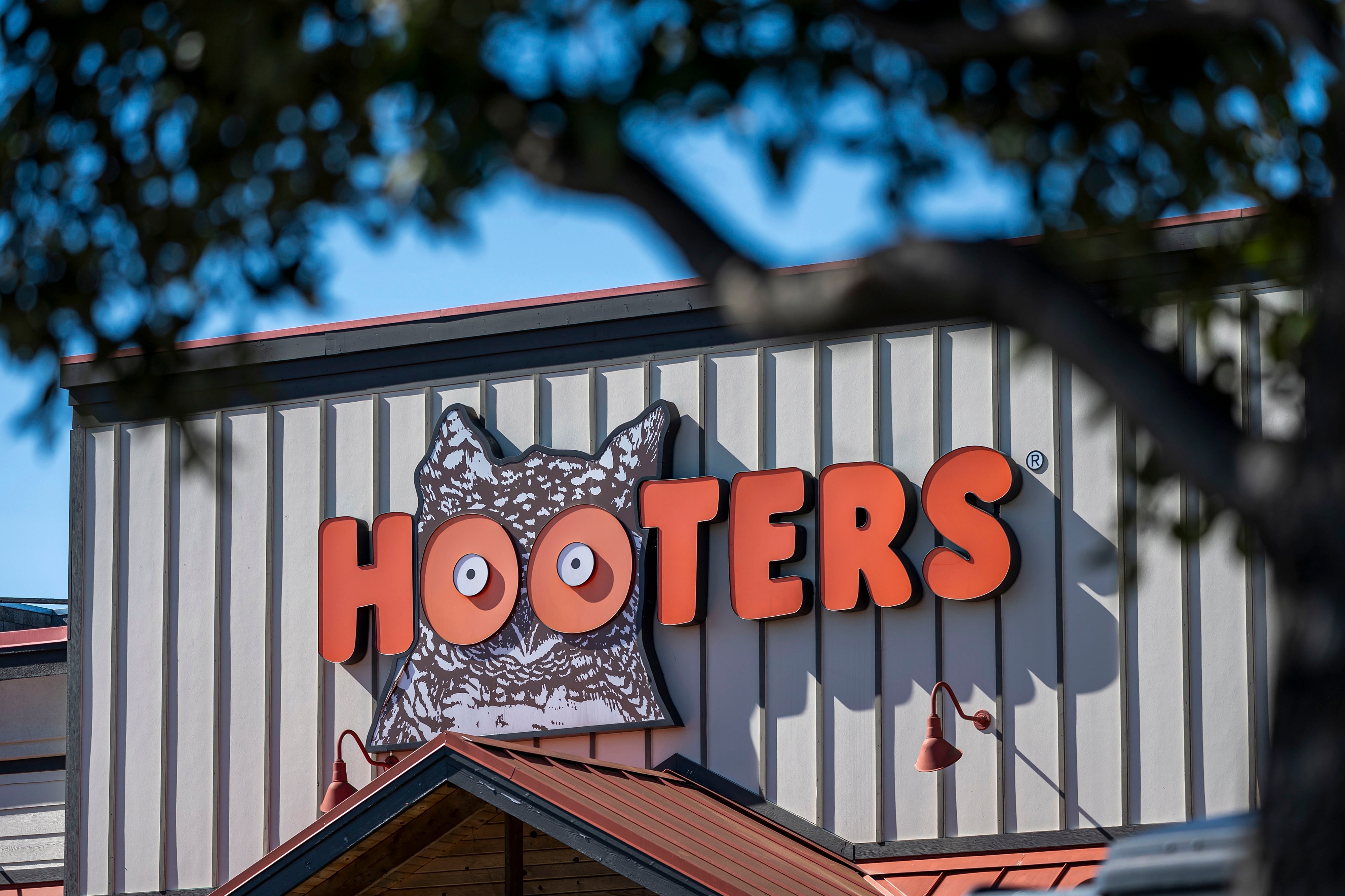 Hooters restaurant exterior with a prominent logo featuring owl eyes on the building facade. Trees partially frame the view