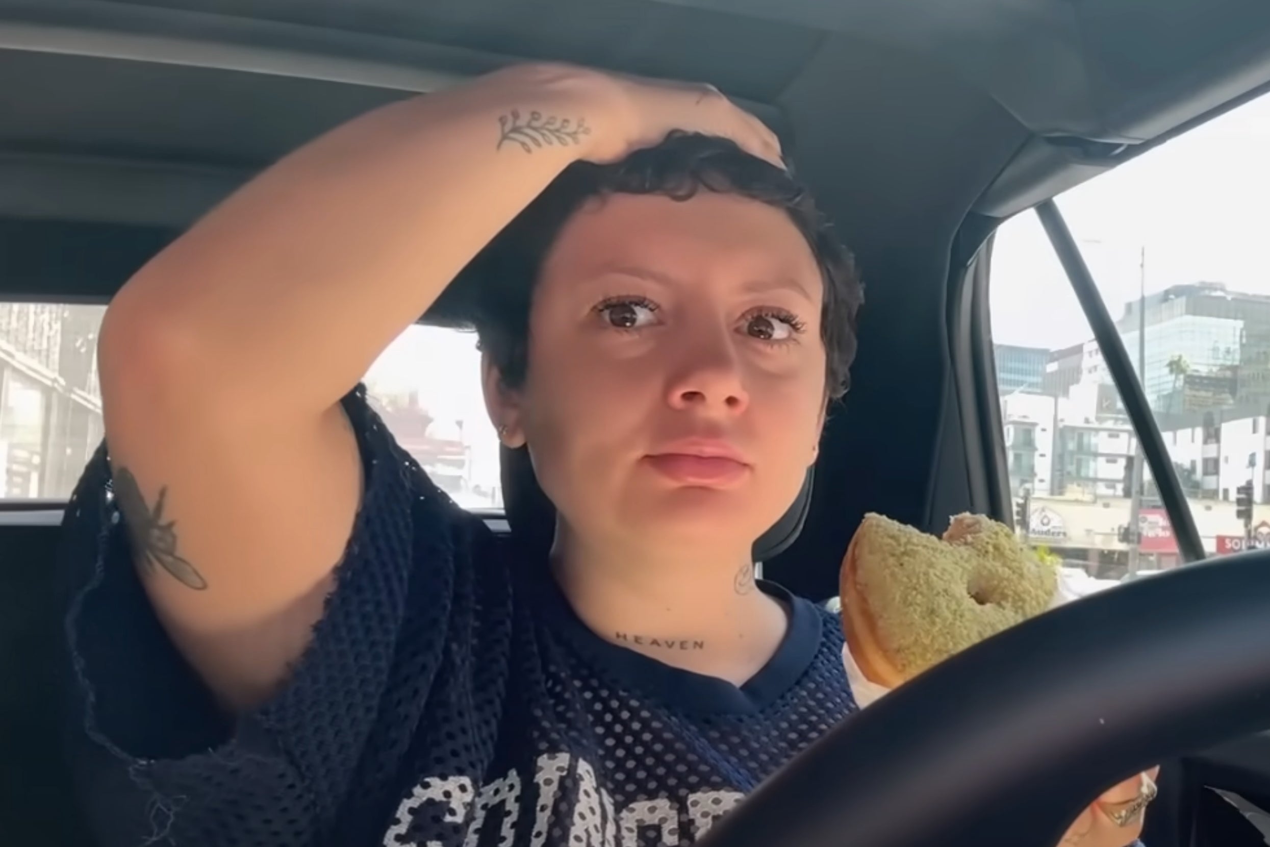 Vanilla Mace in a car holding a donut, looking at the camera with a hand on her head