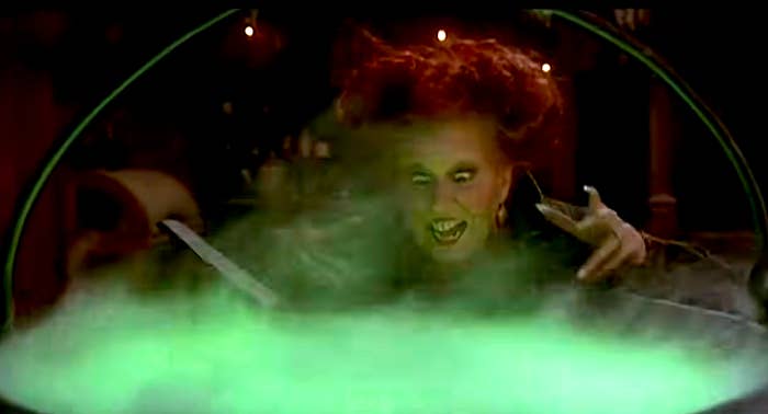 Person with wild hair and pointed nails stirs a glowing green potion in a cauldron, with an intense expression suggesting magic or witchcraft