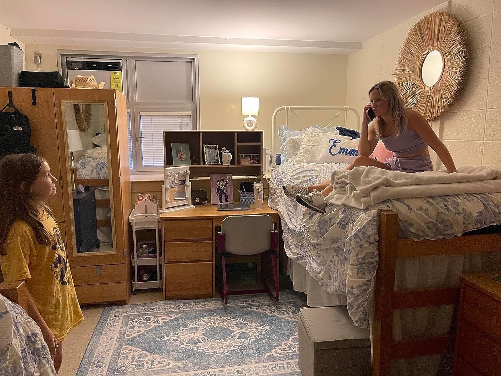 A dorm country   with 2  young people; 1  sits connected  a lofted bed, the different   stands. The country   has organized shelves and a cozy rug