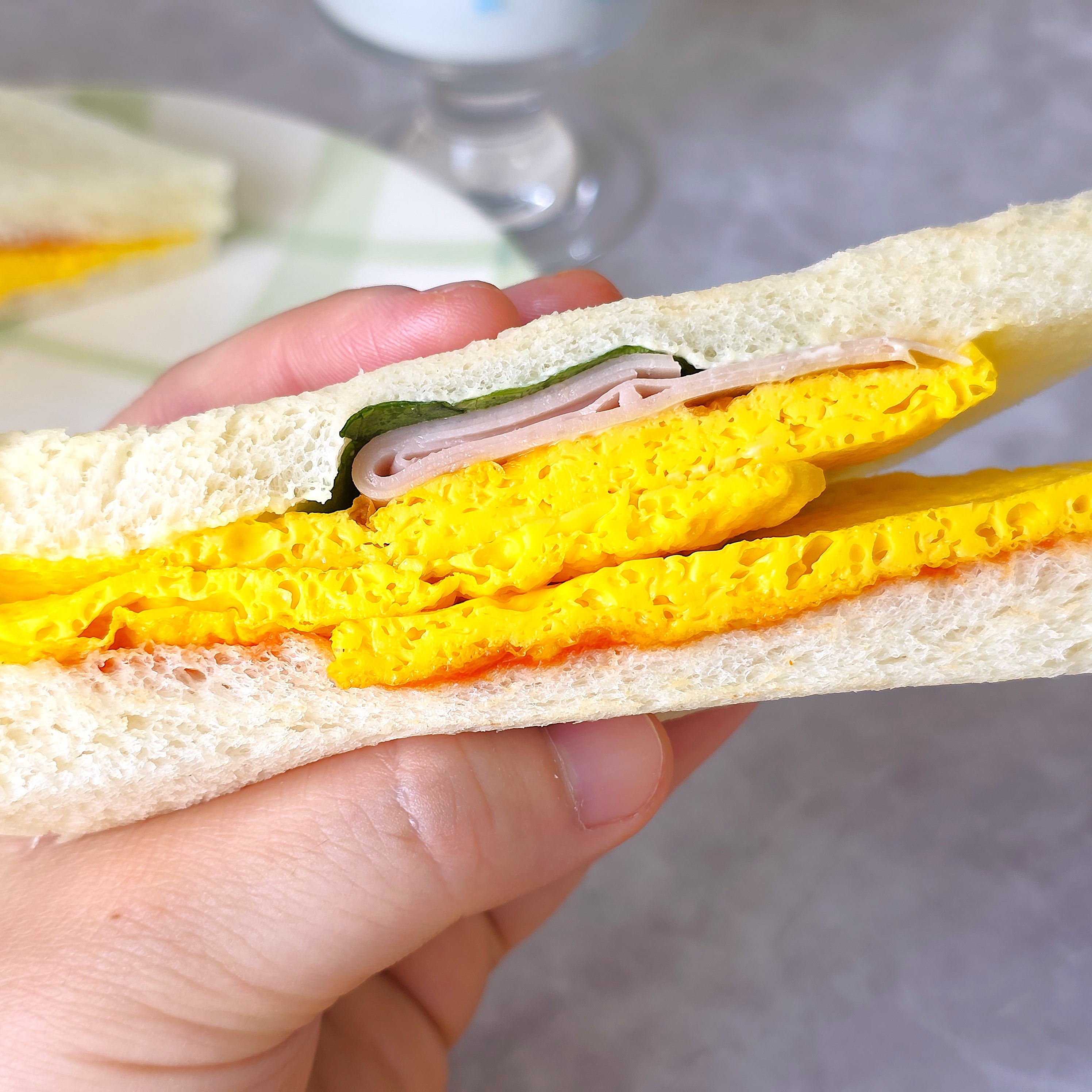 手に持ったサンドイッチの断面。ふわふわの卵とハムが白い食パンに挟まれています。背景は淡い色調です。