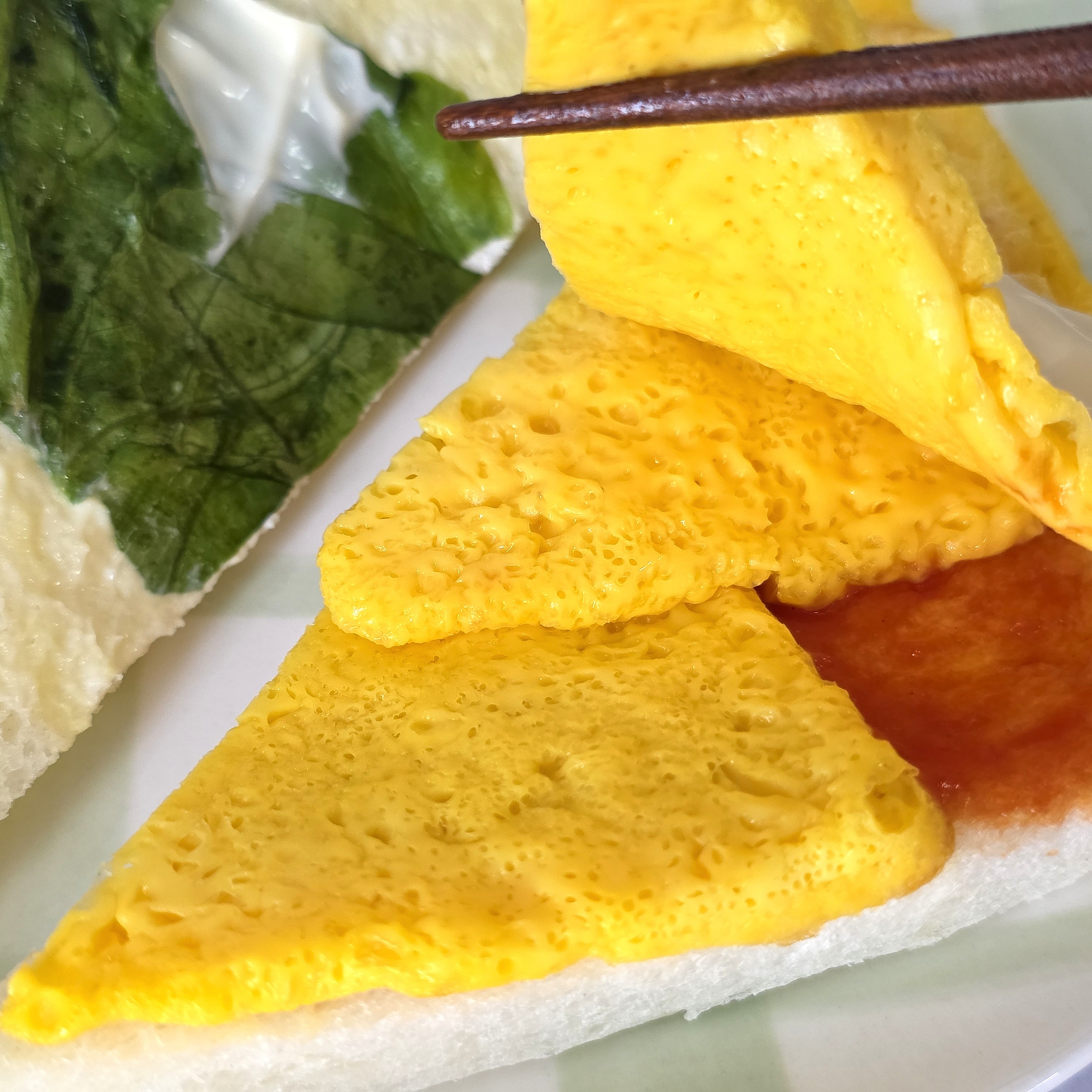 箸で持ち上げた卵焼きがトーストに乗った食事の一部分、ソースと大葉も見える。