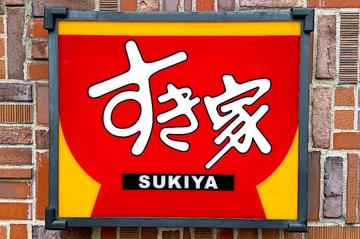 A Sukiya restaurant sign with bold white Japanese characters on a red backdrop, mounted on a brick wall