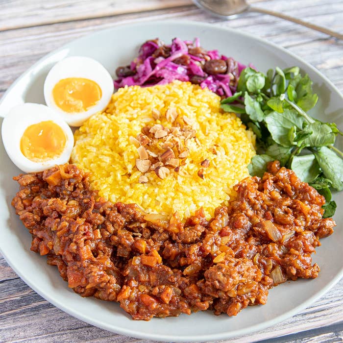 半熟卵、黄色のライス、ドライカレー、ピクルス野菜、緑の葉野菜が並ぶ美しく盛り付けられた料理の写真。
