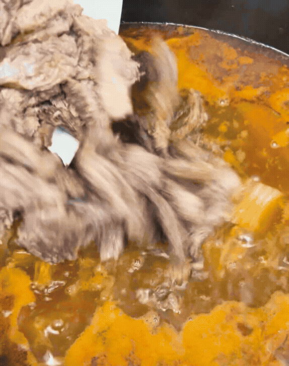 Shredded meat being mixed into a rich, bubbling stew in a pan, creating a hearty dish