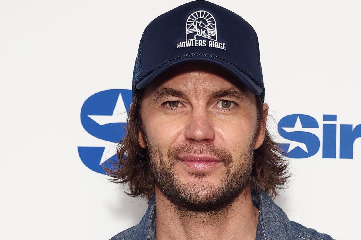 A person wearing a denim button-up shirt and a baseball cap stands in front of a SiriusXM backdrop