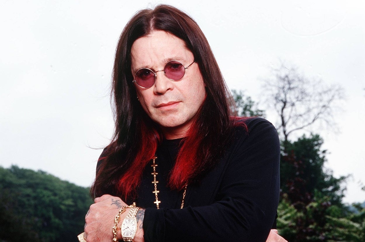 Person with long hair, round sunglasses, wearing a black top and gold jewelry, standing outdoors with arms crossed