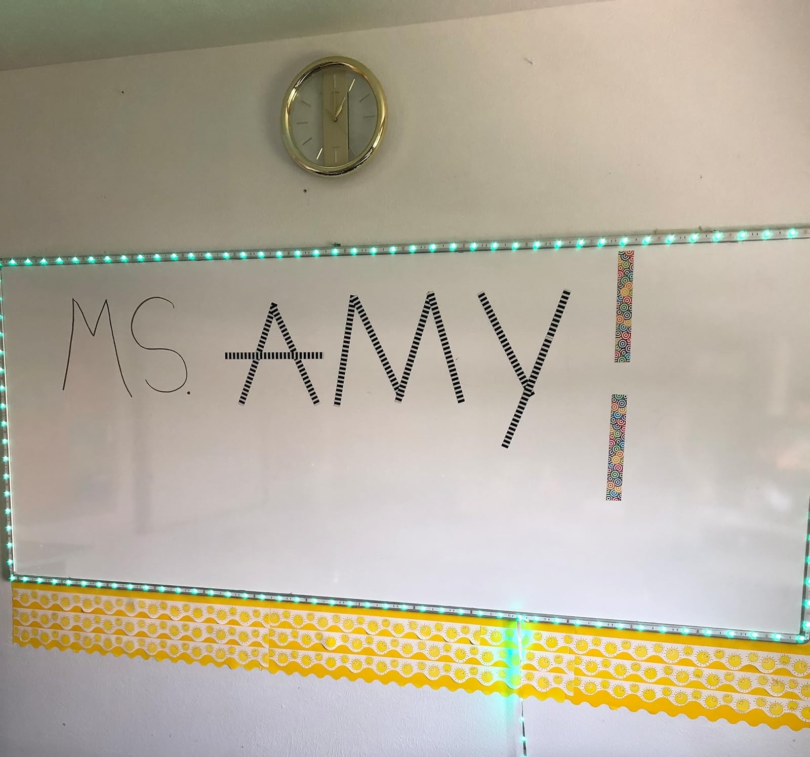 A whiteboard with "Ms. Amy!" written in decorated tape, surrounded by LED lights, with a clock above
