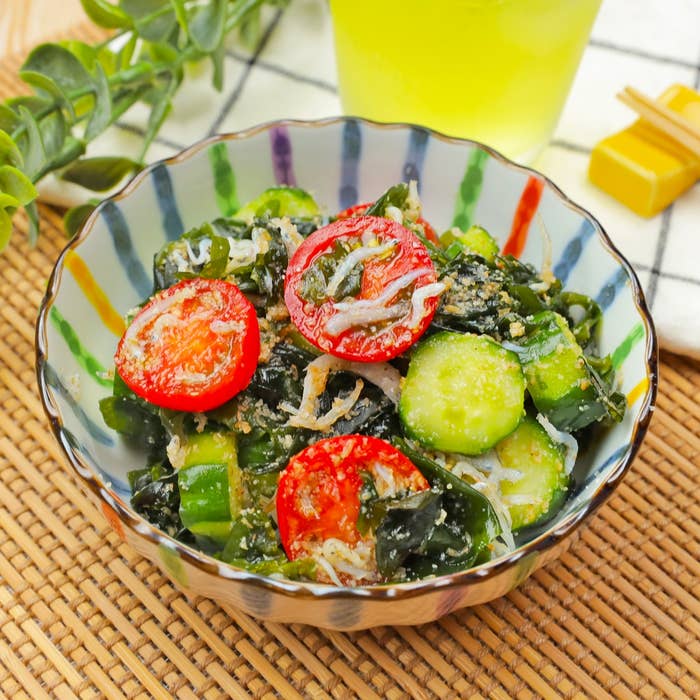 キュウリとトマト、海藻、しらすのサラダが器に盛られている。背景に飲み物と緑の植物がある。