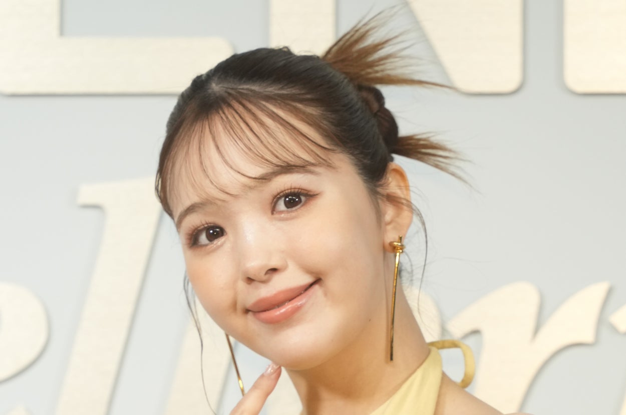 女性が笑顔でポーズを取っている。ノースリーブのドレスを着用し、シンプルなジュエリーを身につけている。背景には文字がある。