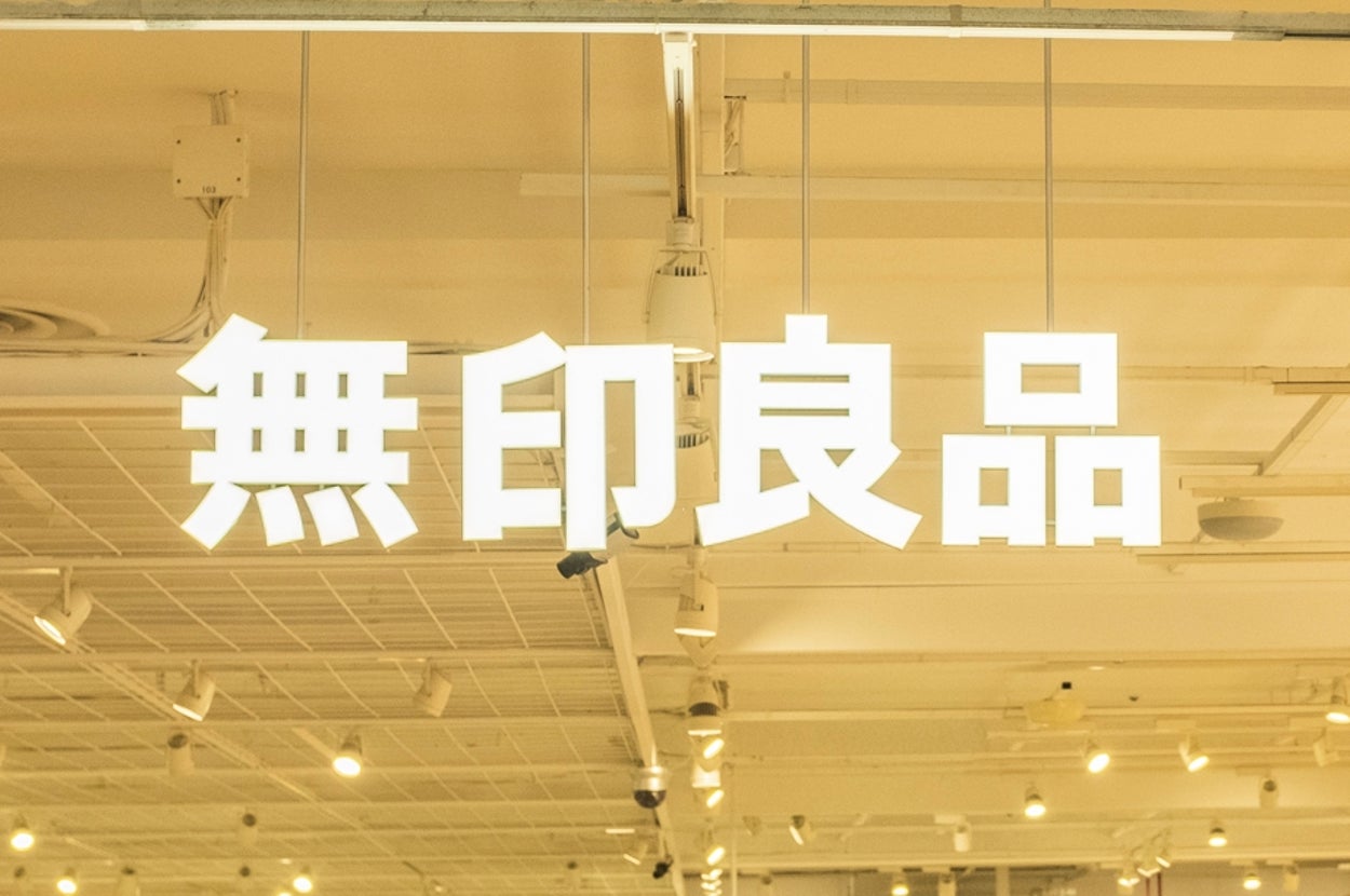 People shopping in a well-lit, spacious Muji store with organized shelves and displays. Japanese text over the entrance