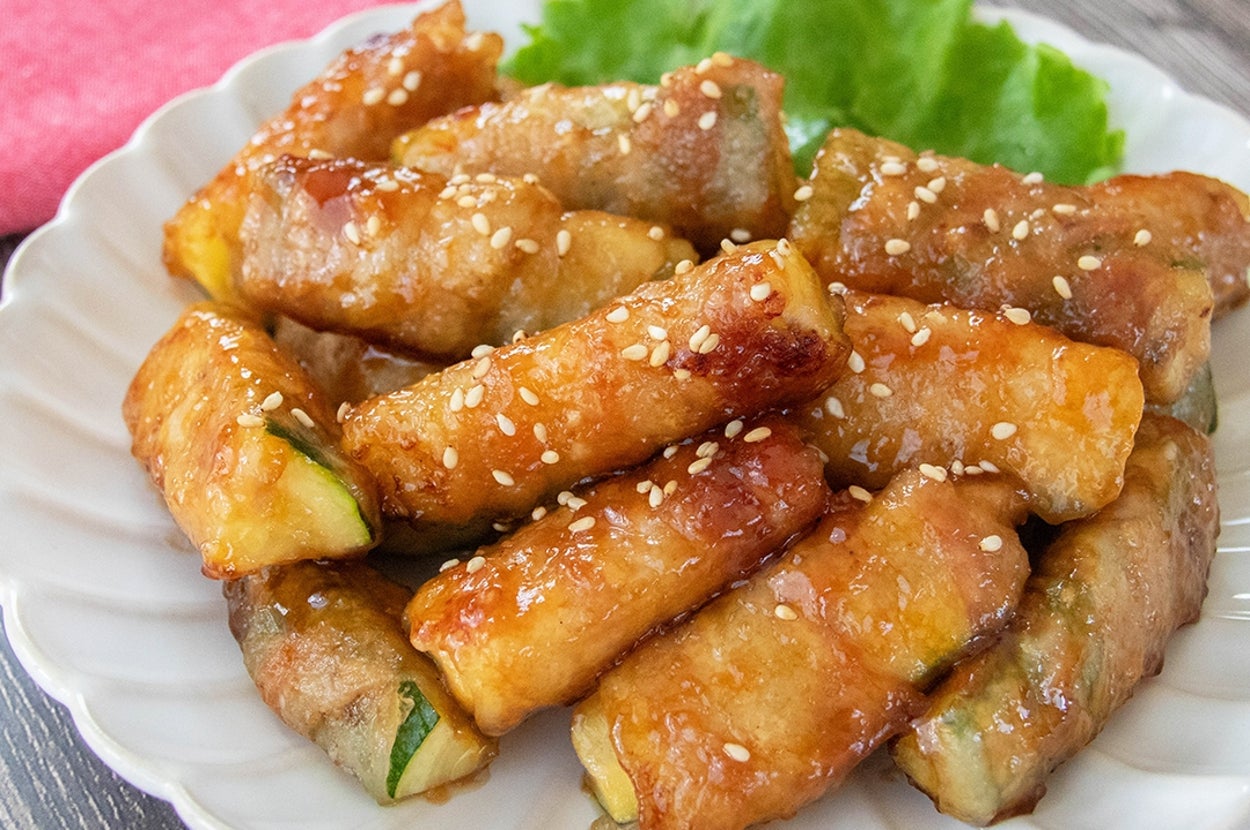 お皿に盛られた肉巻きズッキーニ。白いりごまがトッピングされ、美味しそうに焼き上がっている。バックにレタスが添えられている。