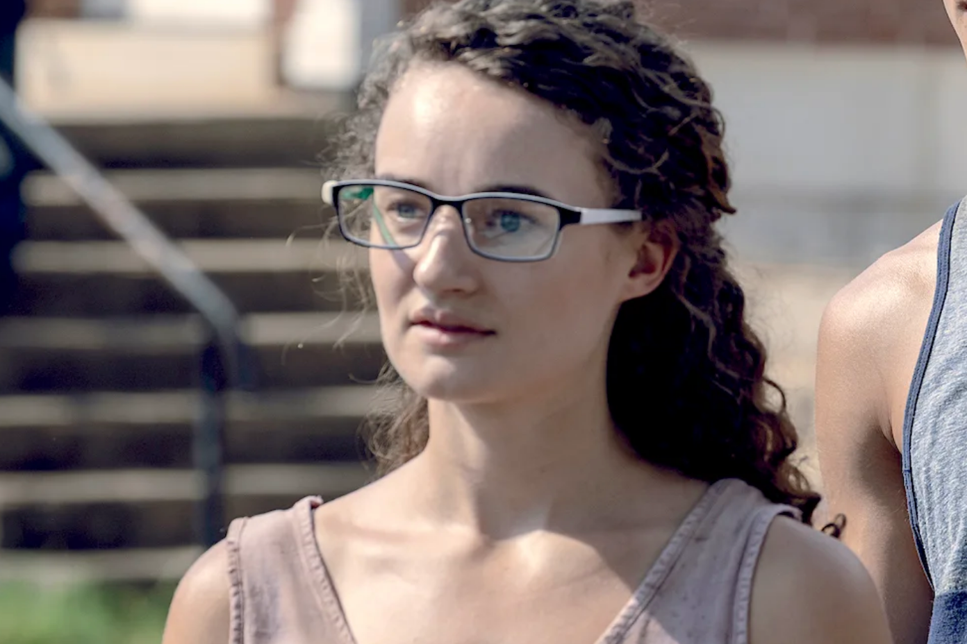 Person with curly hair and glasses, wearing a sleeveless top, looks forward in an outdoor setting