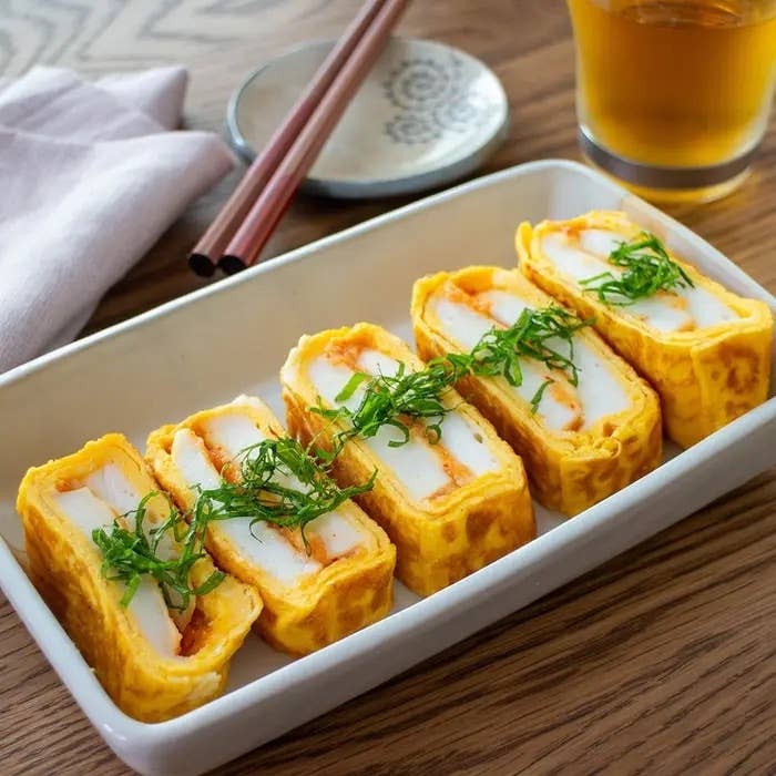 長方形のお皿にのった厚焼き玉子寿司、上に細切りの青ネギがのっている。背景にはお茶と箸が見える。