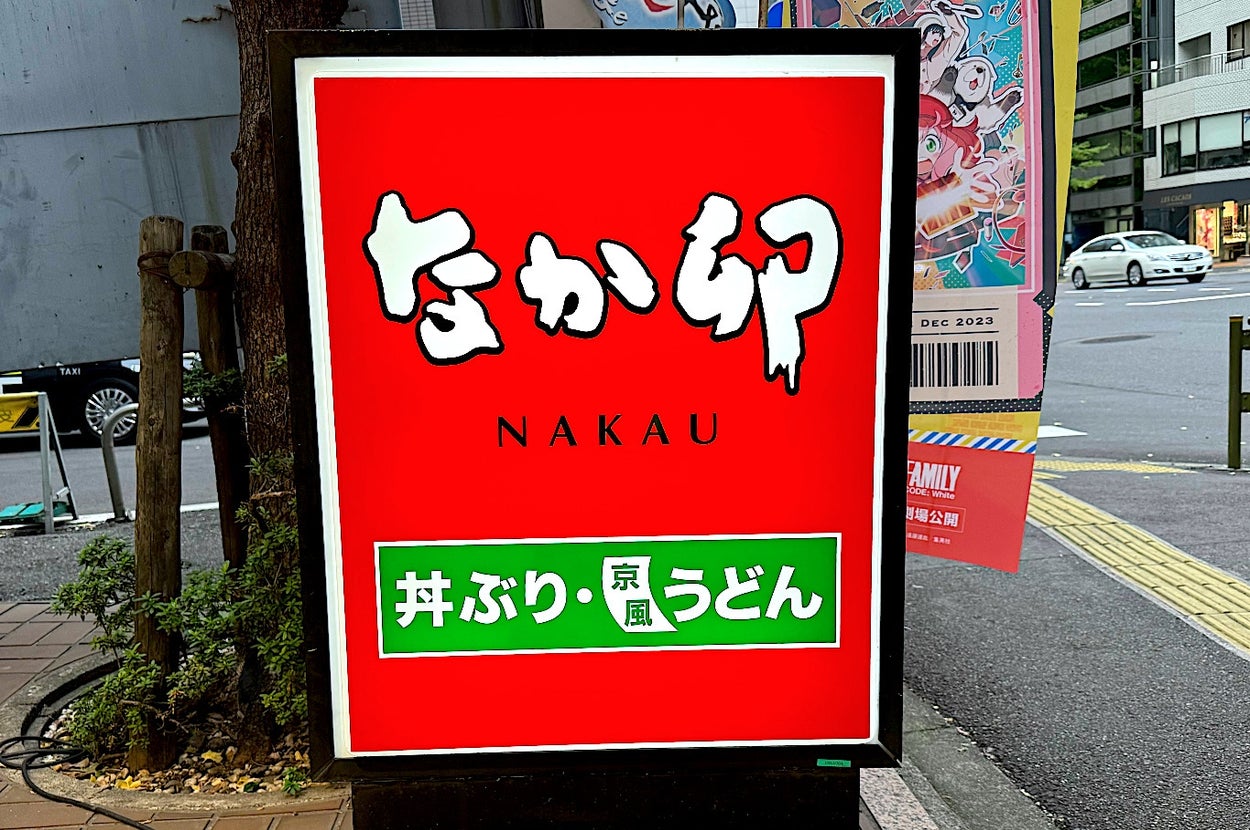 A street sign for "Nakau" with Japanese text and illustrations, promoting menu items like donburi and udon, next to a sidewalk and city street