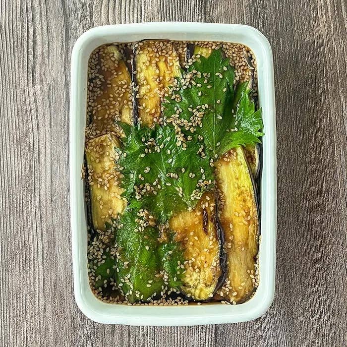 長方形の白い皿に盛り付けられた茄子の炒め料理。上にゴマと新鮮な葉がトッピングされています。