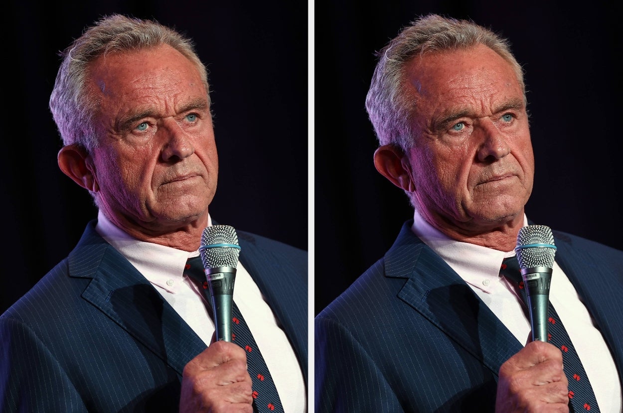 A man in a suit and tie holds a microphone, appearing to address an audience at an event