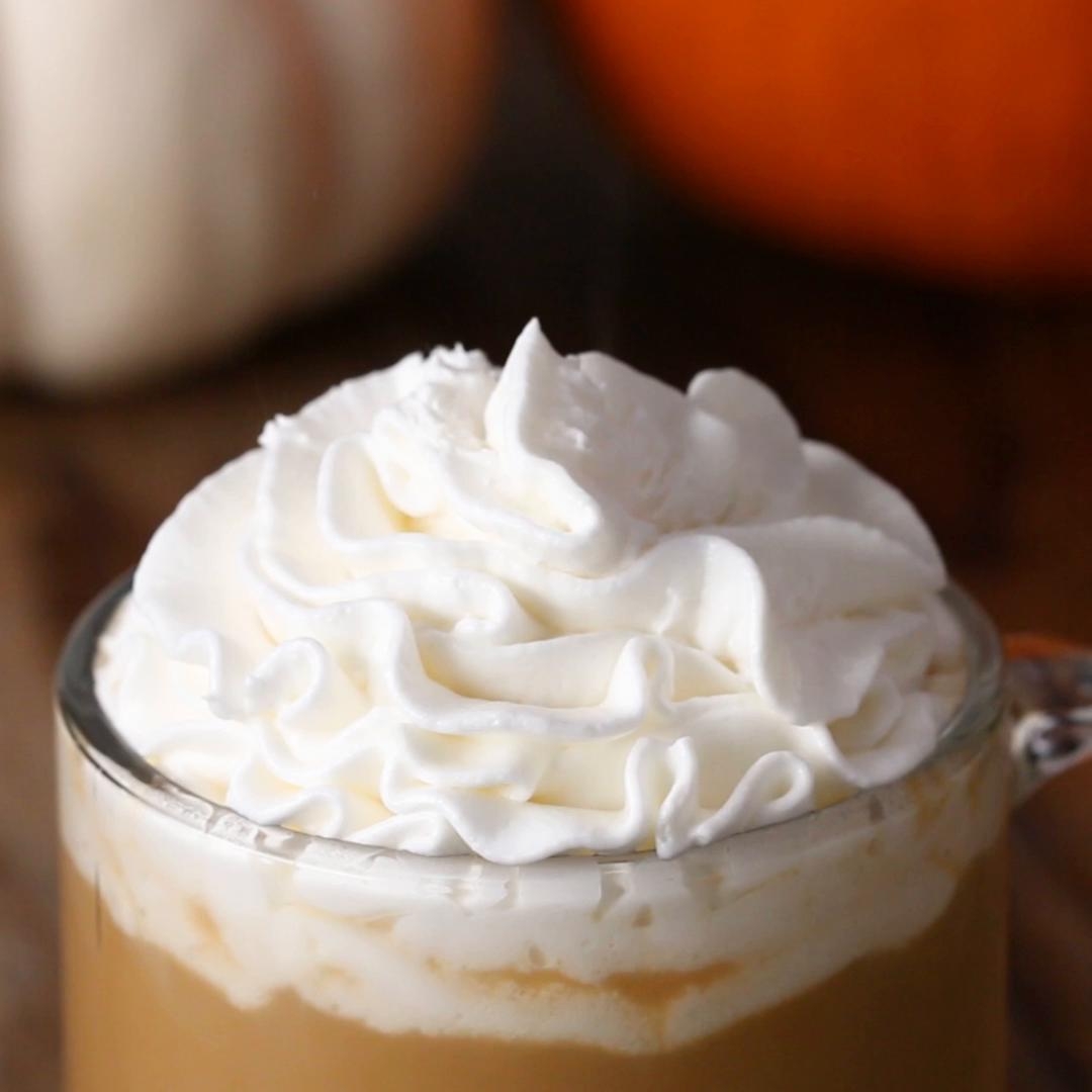 A glass mug filled with creamy coffee topped with a generous swirl of whipped cream, with a pumpkin blurred in the background