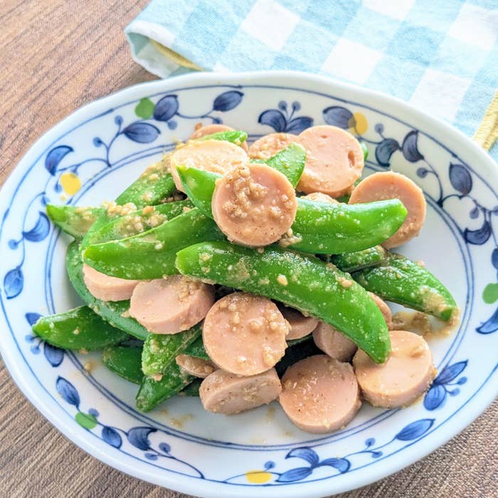 スナップエンドウと魚肉ソーセージの炒め物が皿に盛られています。背景にチェック柄のナプキンがあります。