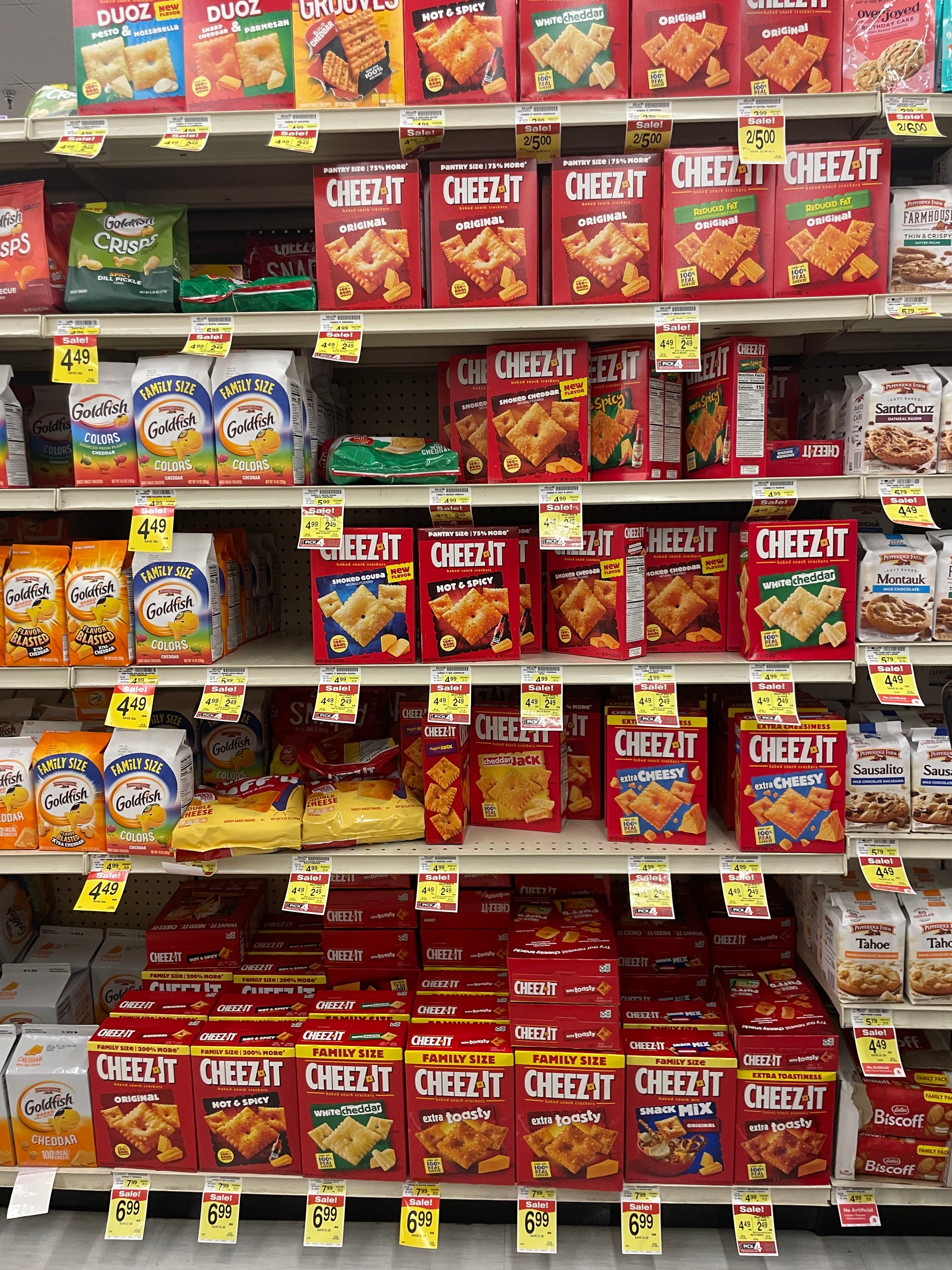 Store shelf with various Cheez-It cracker boxes. Nearby shelves contain crackers and snacks from other brands