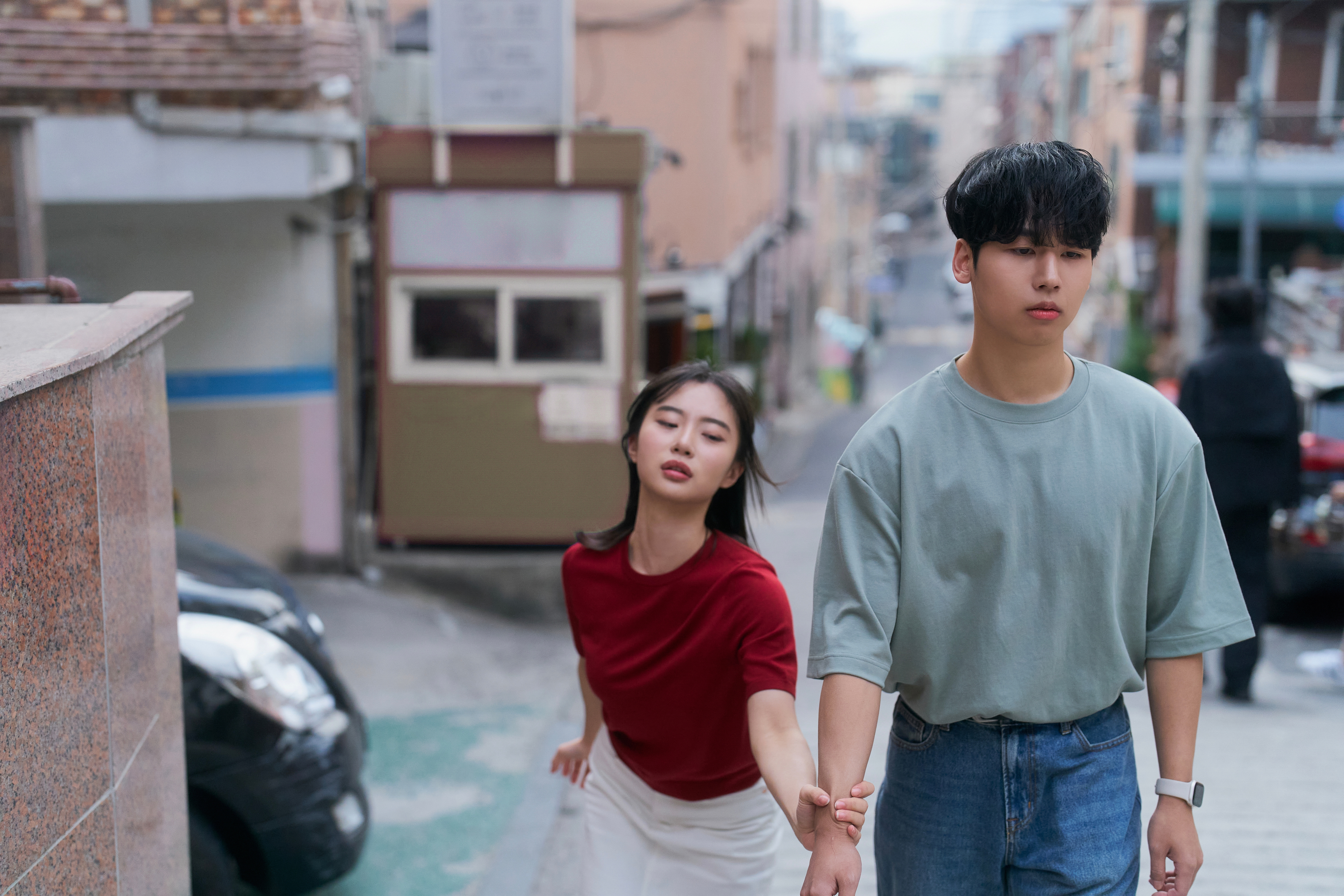 A person in a red shirt reaches out to another person in a casual outfit on a narrow urban street
