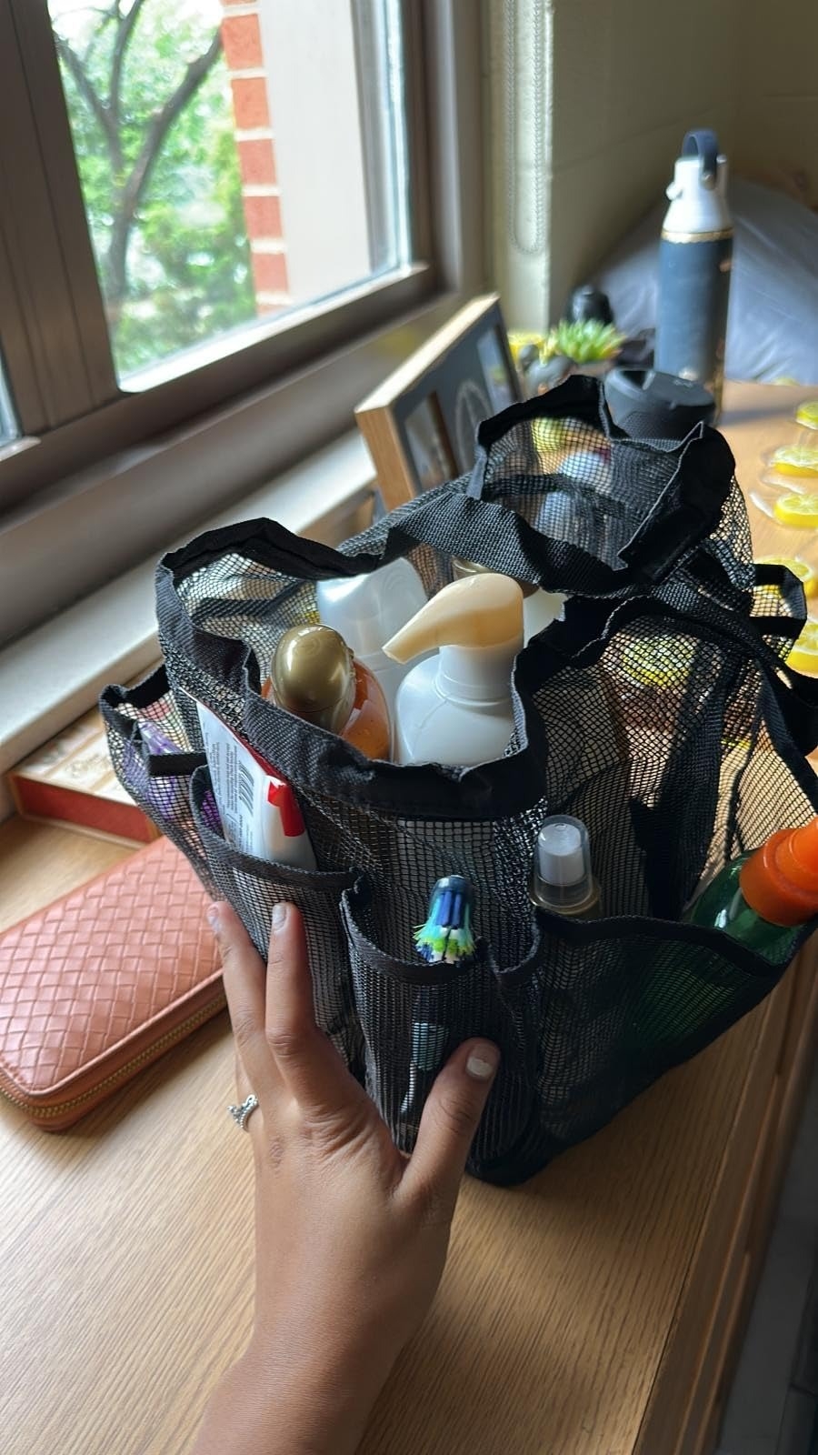 Mesh toiletry organizer filled with various bottles and tubes, held by a person with manicured nails, placed on a wooden surface near a window