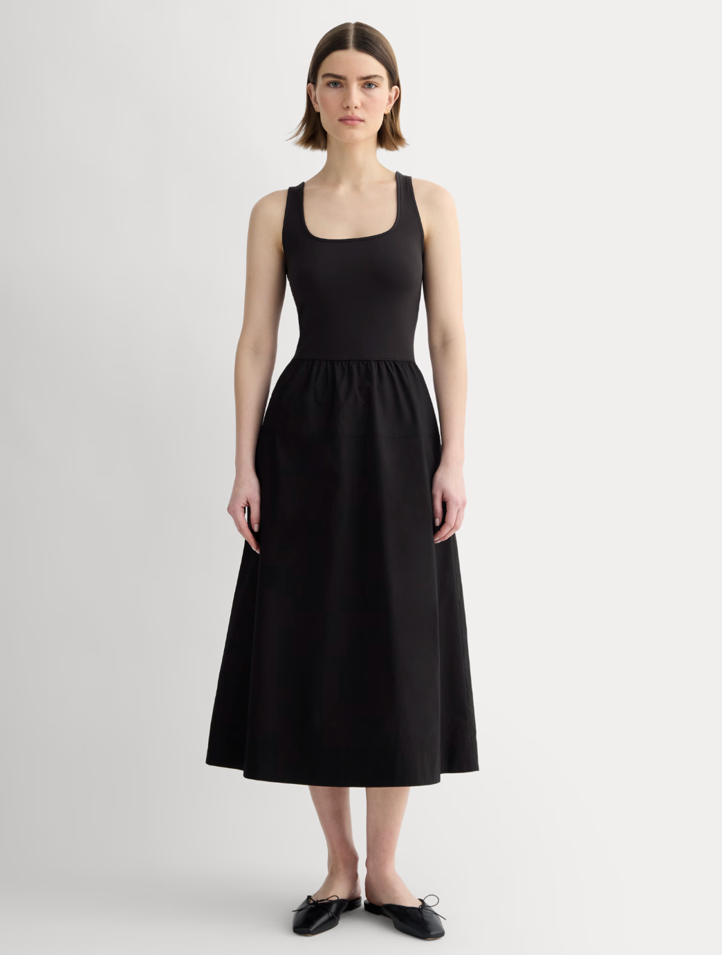 Person wearing a sleeveless black dress and black ballet flats poses against a plain background