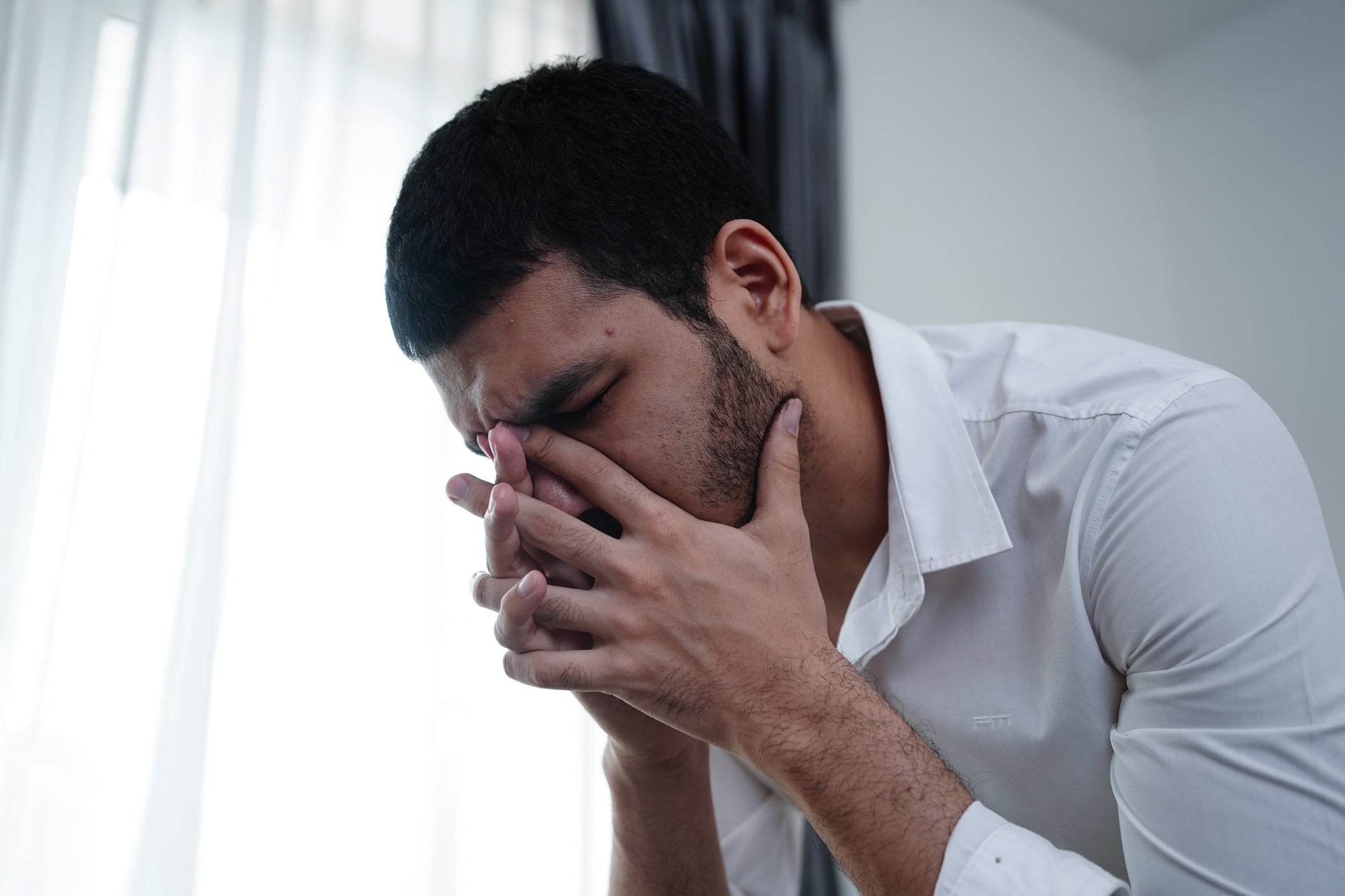 Person i en hvit skjorte med hendene på ansiktet, virker stresset eller kontemplativ, stående innendørs i nærheten av et vindu med gardiner