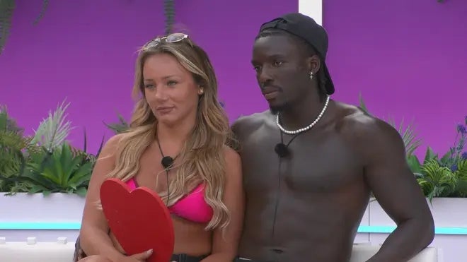 Two individuals posing together, one holding a red heart prop. The man is shirtless with a cap, and the woman is wearing a bikini top with sunglasses on her head