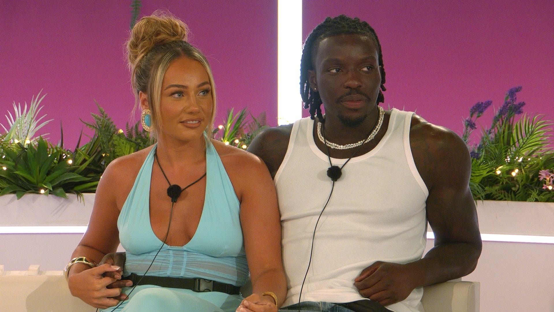 Two people seated, wearing casual attire with microphone packs, likely on a TV show set. The woman in a sleeveless outfit, the man in a tank top