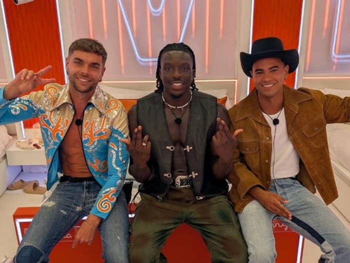 Three people sitting on a red-carpeted set; two wearing Western-style jackets, one in a sheer top and leather pants