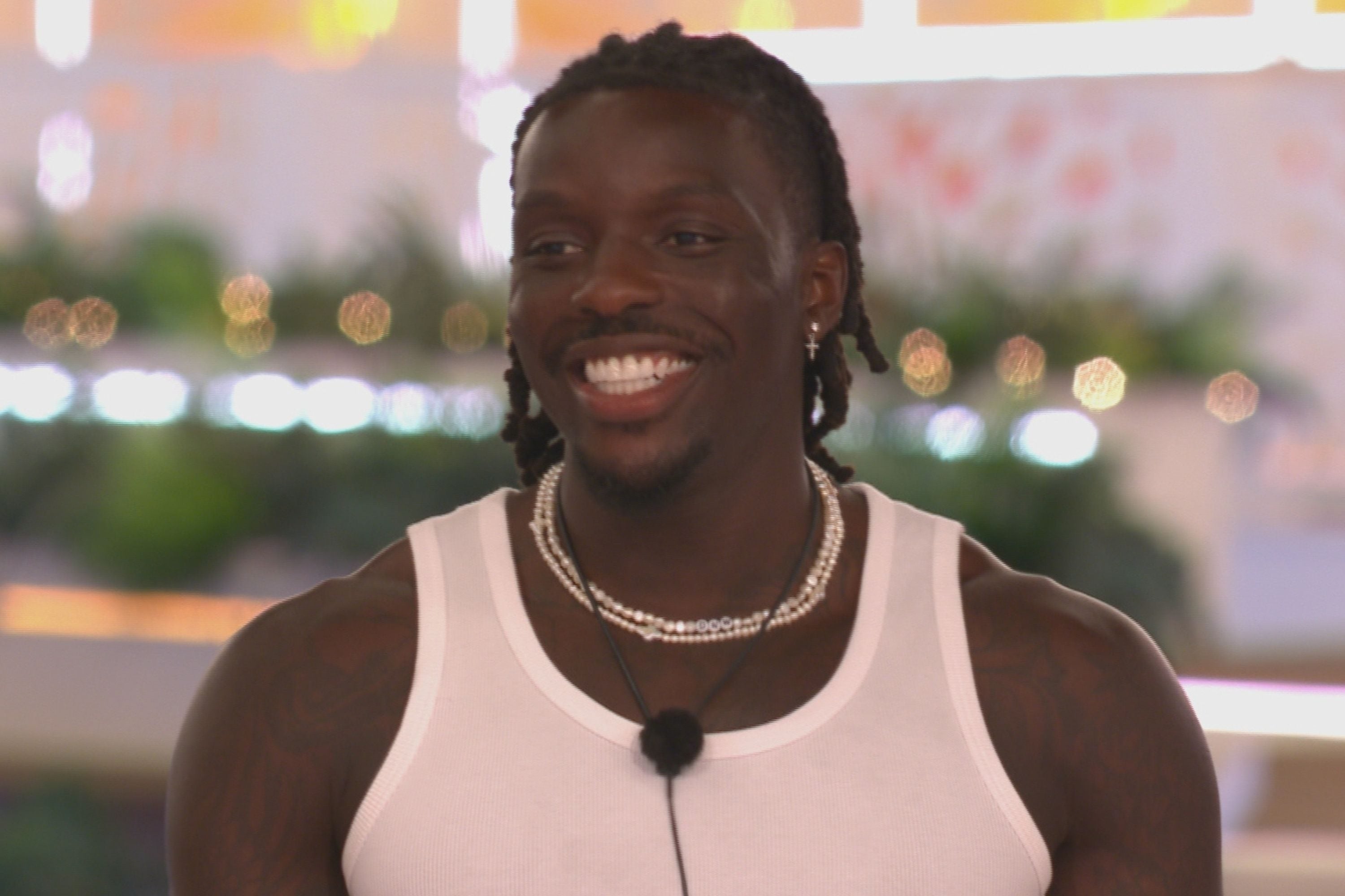 Man wearing a white tank top and pearl necklaces, smiling and standing in a lively setting
