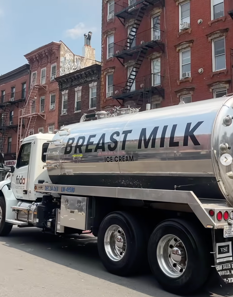 Tanker truck labeled "Breast Milk Ice Cream" parked on a city street with brick buildings in the background