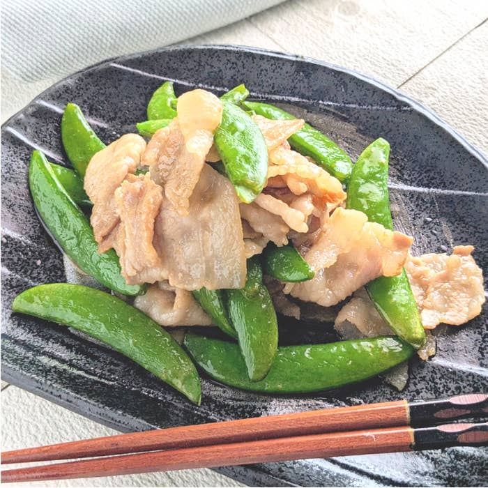 スナップエンドウと豚ばら肉の炒め物が黒い皿に盛り付けられています。箸がそばに置かれています。