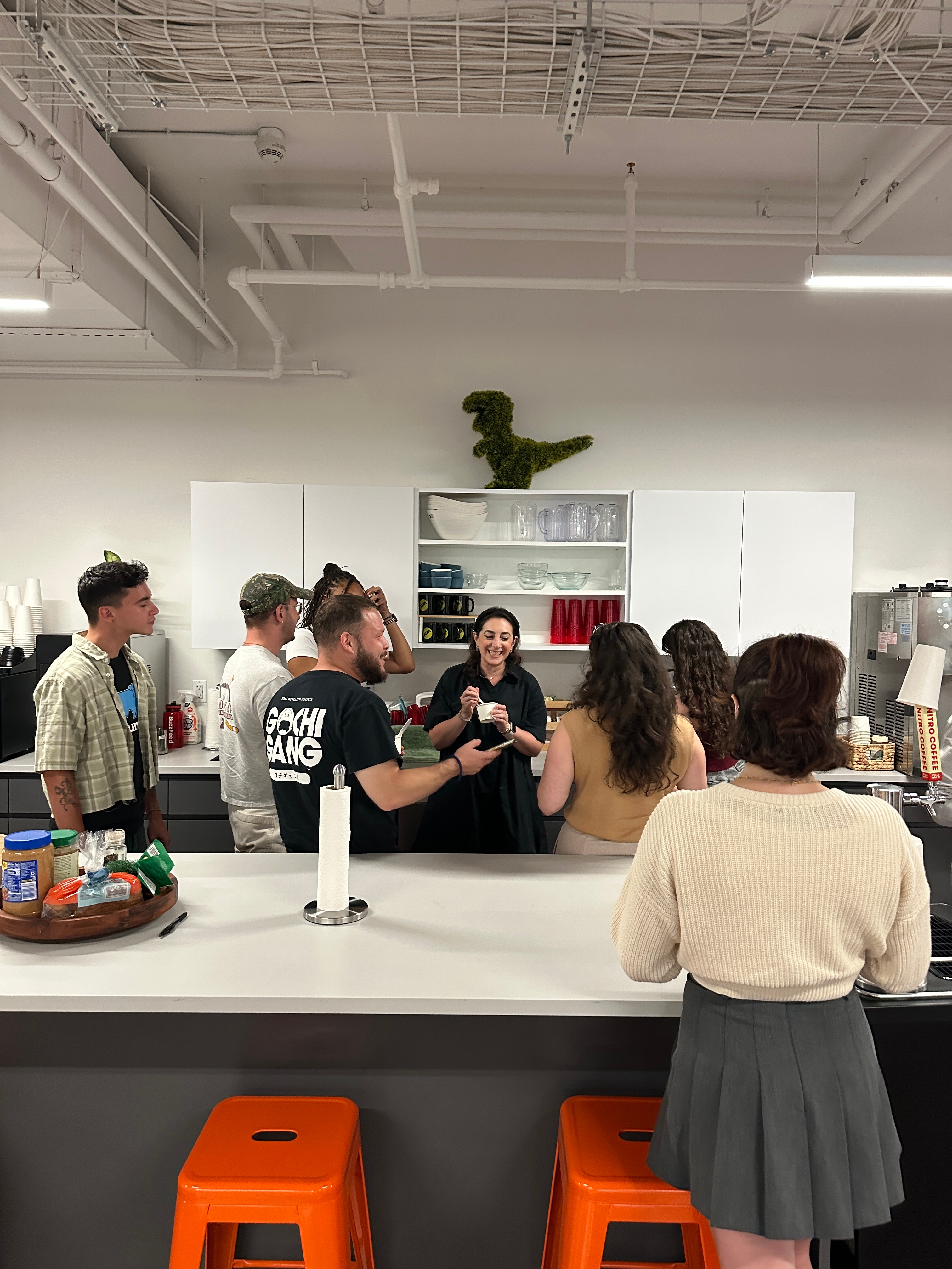People gathered around a kitchen island chatting and sharing drinks in a modern breakroom setting, with snacks and a dinosaur decoration visible