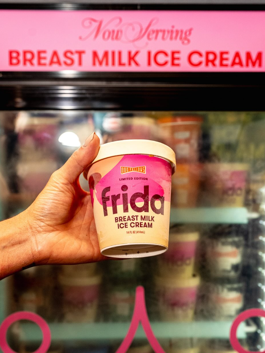 Hand holding a "Frida" branded ice cream tub labeled as breast milk ice cream, with a freezer display in the background