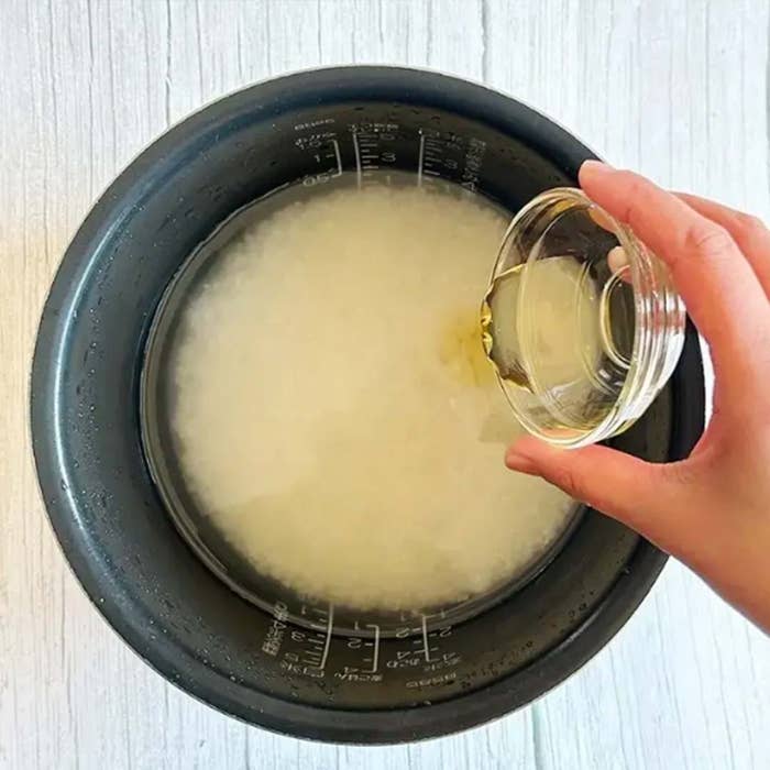 炊飯器の中で水に浸された米に液体を注ぐ手。