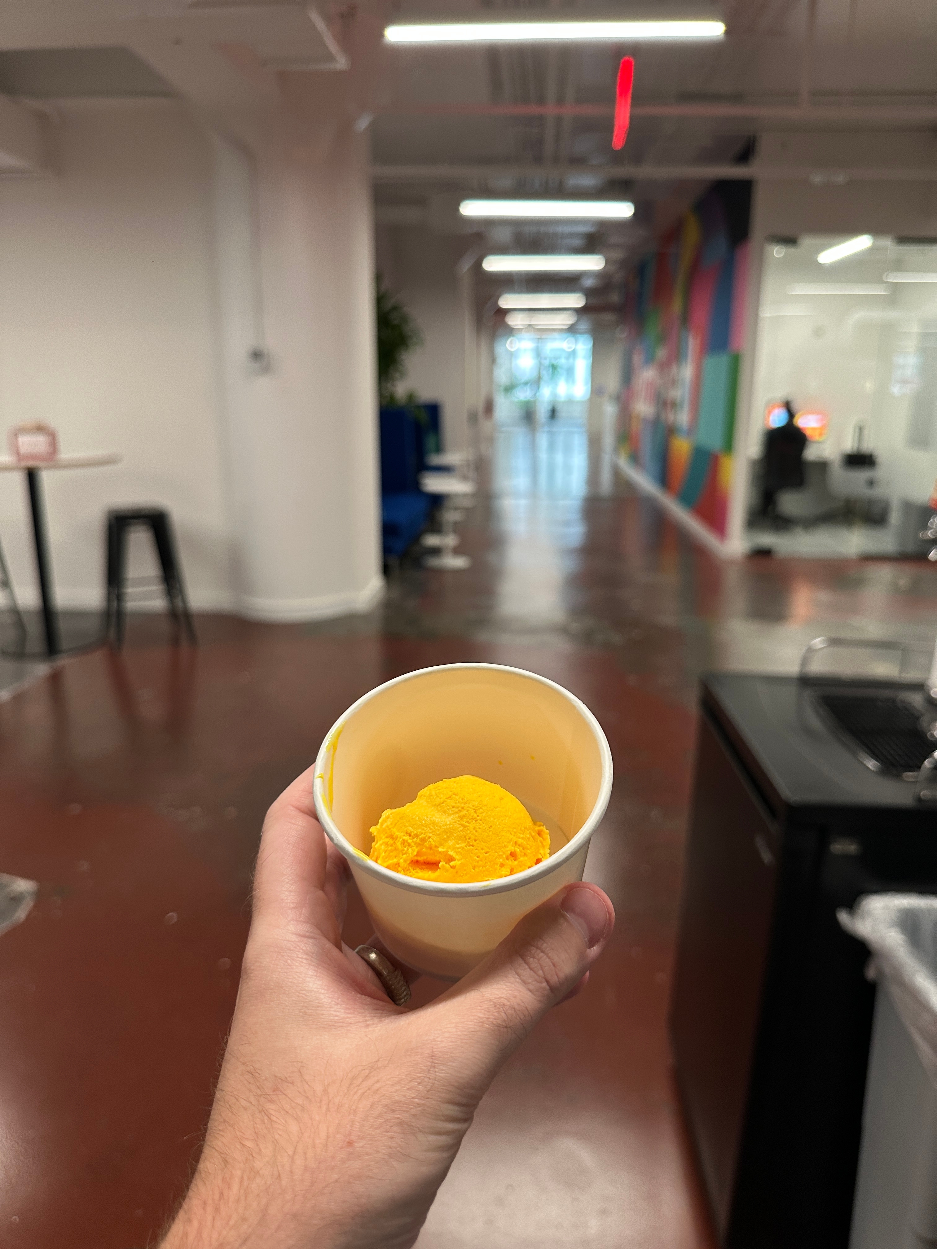 Hand holding a cup of yellow ice cream in a modern office hallway