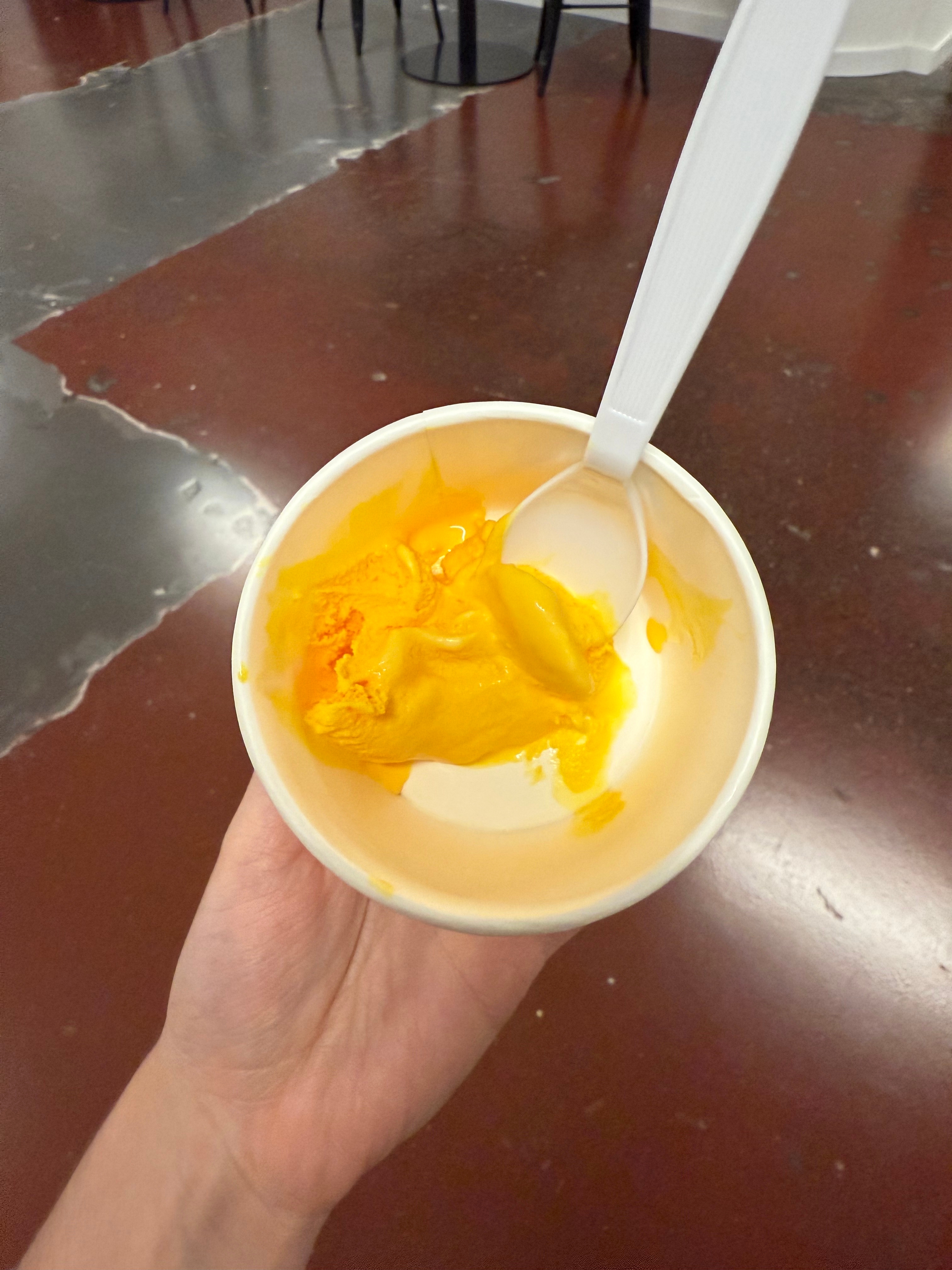 A hand holds a mostly empty cup of mango gelato with a white plastic spoon indoors