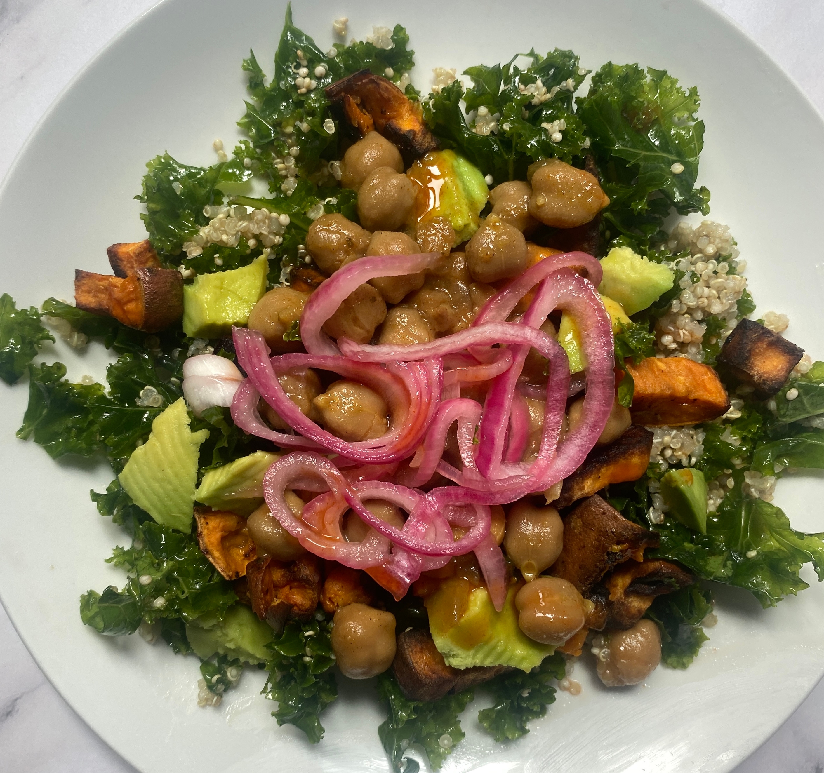 A plate of salad with kale, avocado slices, chickpeas, quinoa, roasted sweet potatoes, and pickled onions