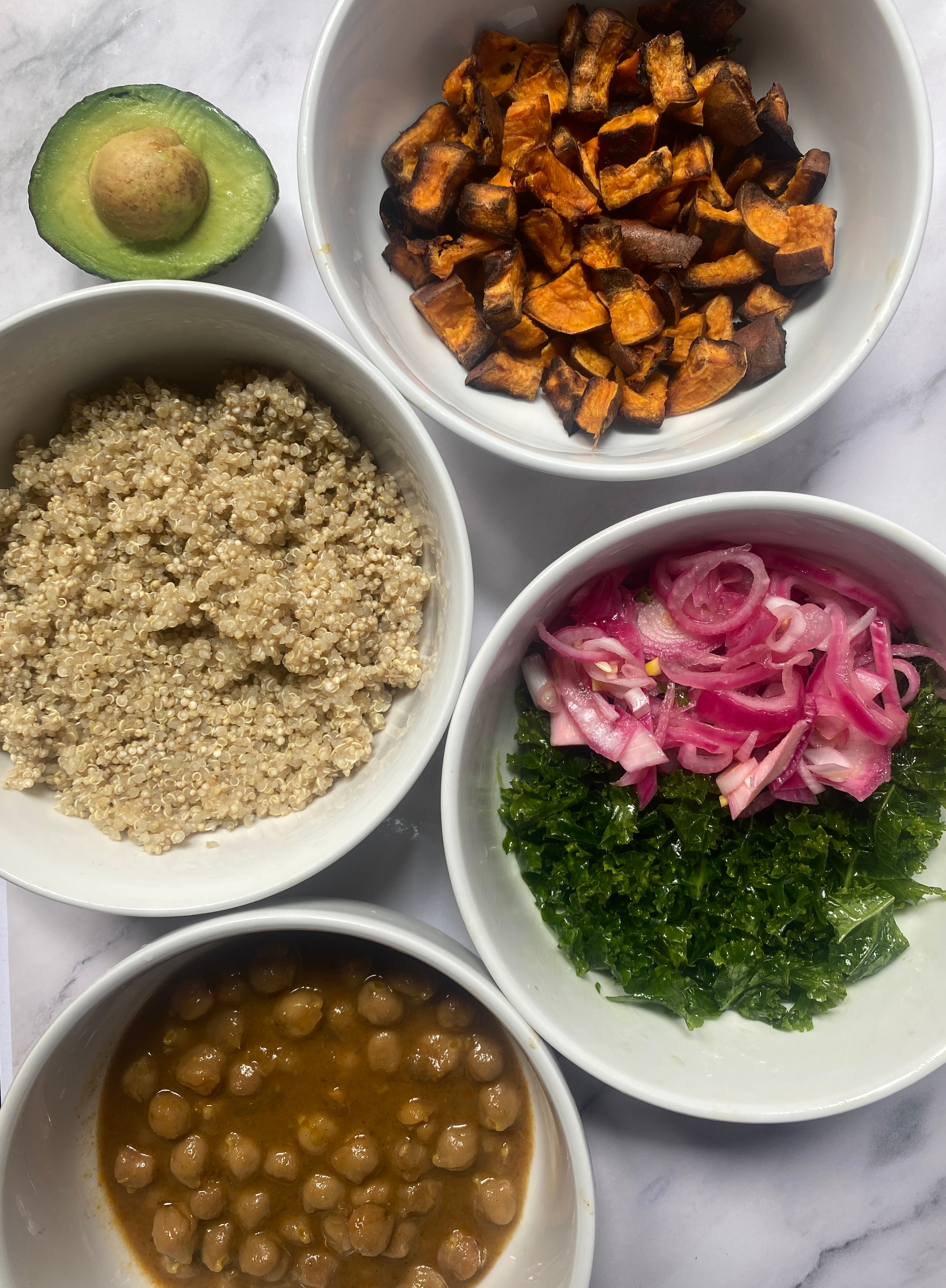 Bowl with quinoa, roasted sweet potatoes, pickled onions, kale, and chickpeas. Half avocado placed nearby on a marble surface
