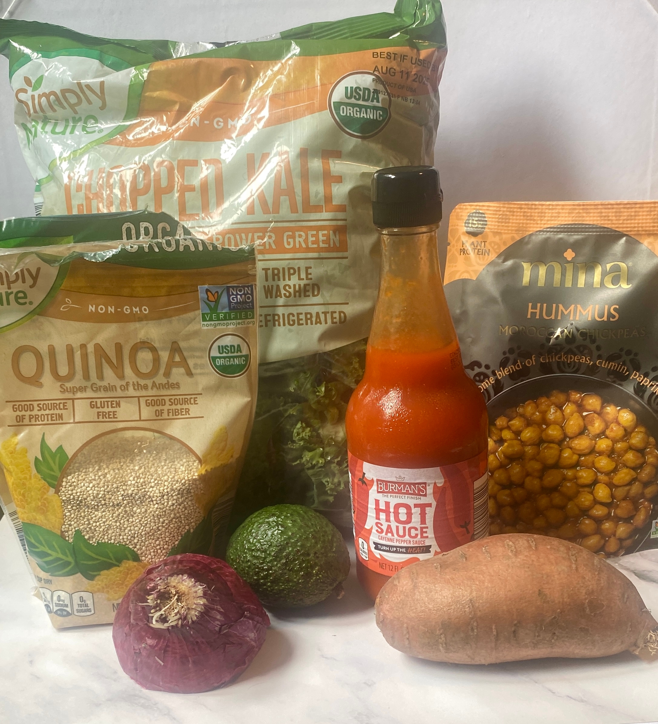Organic food items including kale, quinoa, hot sauce, hummus, avocado, red onion, and sweet potato are displayed on a table