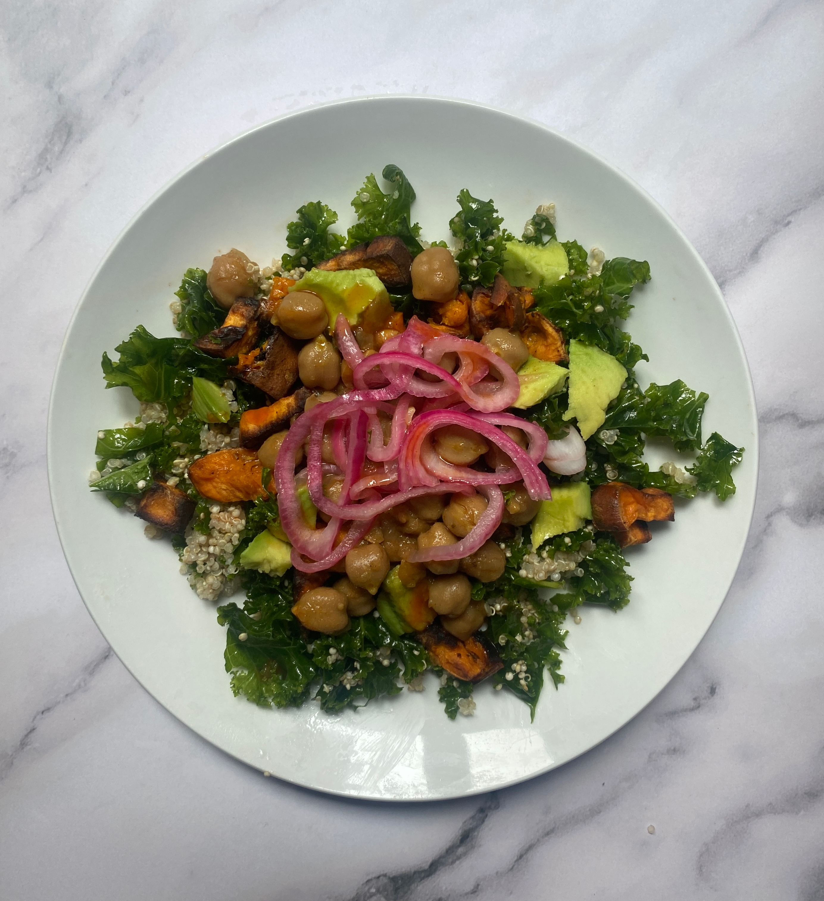 Plate of kale salad with quinoa, chickpeas, avocado, roasted sweet potatoes, and pickled onions on a marble surface