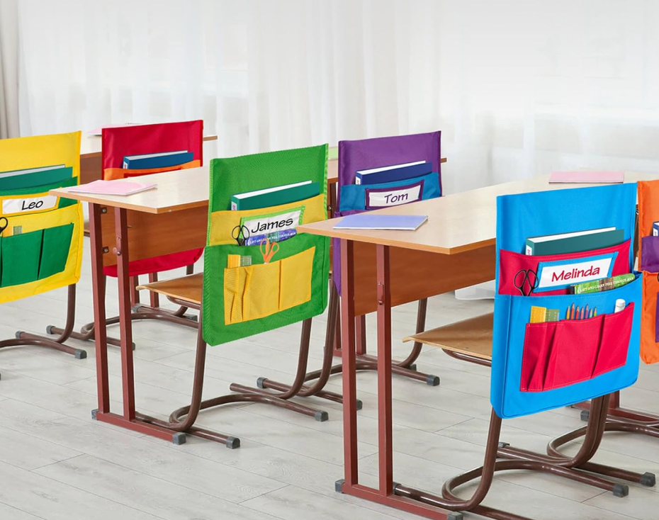 Classroom desks with colorful chair pockets holding books and supplies, each labeled with a student's name