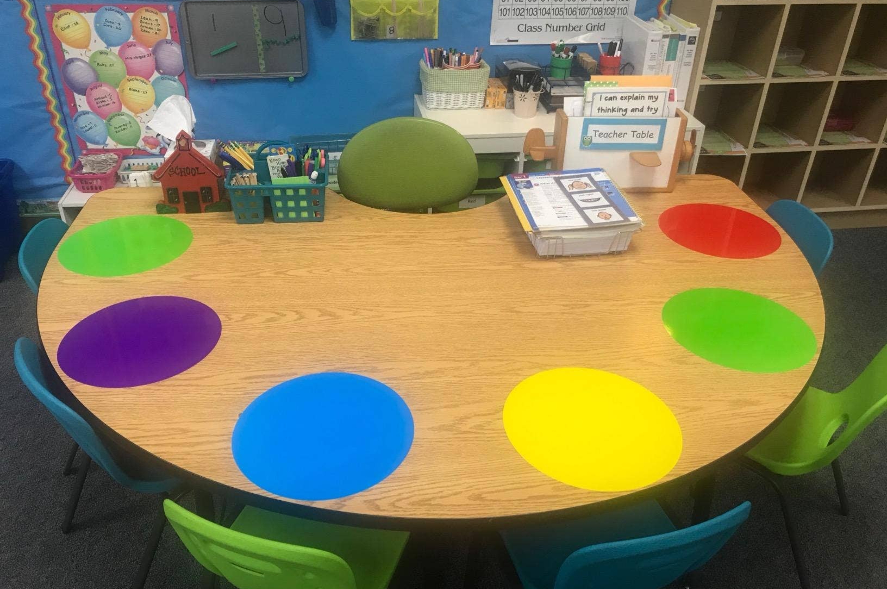 Colorful classroom table with labeled bins and books, surrounded by chairs. The table is set up for organized student activities