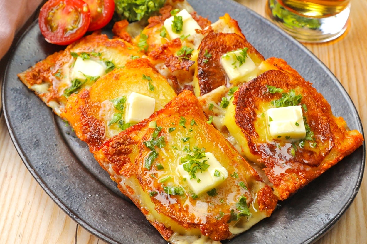 お皿に盛られたチーズとバターをのせたこんがり焼けたポテトパンケーキ。横にトマトスライスと緑の付け合わせが添えられています。